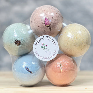 A set of six colorful bath bombs in clear packaging labeled “Island Thyme Bath Bomb Sampler,” each with a dried flower or herb on top, displayed on a wooden surface.