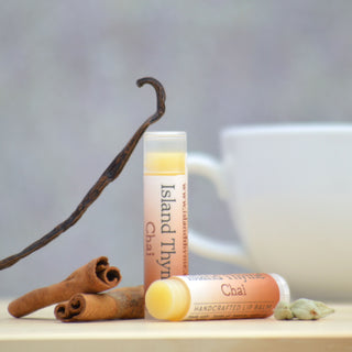 Two tubes of Chai Lip Balm are shown on a wooden surface with cinnamon sticks, vanilla bean, cardamom pods, and a white cup in the background.