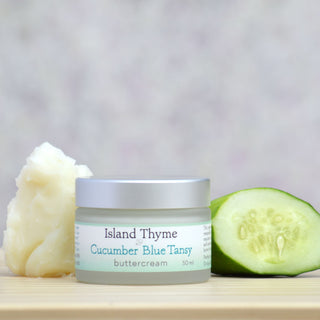 A jar of Cucumber Blue Tansy Buttercream, ideal as a night cream for face care, rests on wood beside a dollop of cream and cucumber slices against a soft, neutral background.