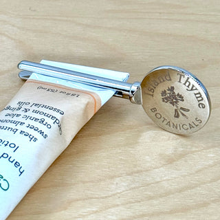 The Lotion Key - Travel Lotion Tube Squeezer holds a travel lotion tube on a wooden surface. Its round end is engraved with “Island Thyme BOTANICALS” and features an illustration of a plant.