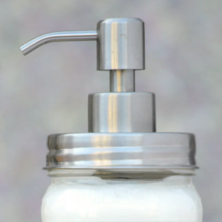 A close-up of the Eco – lotion Pump, a 16oz dispenser fitted on a glass jar partially filled with white lotion, against a blurred background.