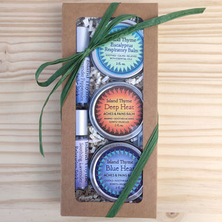 The Remedies & Soothers Essentials gift box includes three Island Thyme balm tins—respiratory, analgesic, and another balm—plus two lavender lip balms, all tied with a green ribbon and arranged on shredded paper in a brown box.