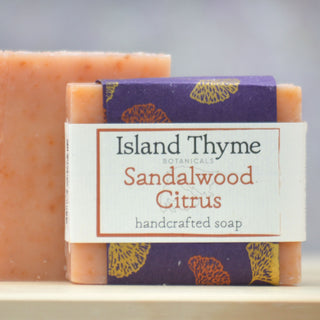 A bar of Sandalwood Soap, orange with specks, sits next to an unwrapped one. Infused with sandalwood and orange oil, it features a purple and cream label with yellow accents.