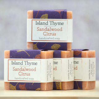 Four bars of Sandalwood Soap are stacked on a light surface, featuring purple and cream labels with orange text and botanical designs—a great shaving soap option with notes of sandalwood and orange oil.