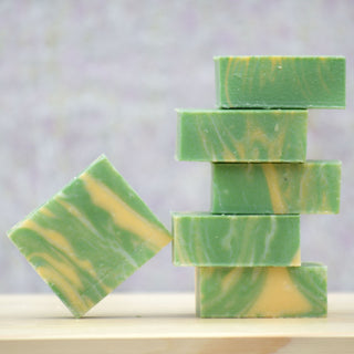 Five Spruce & Cedar Soap bars, green and yellow marbled, are stacked on a wooden surface with one leaning diagonally. Infused with PNW forest scent, the softly blurred background features pastel shades.