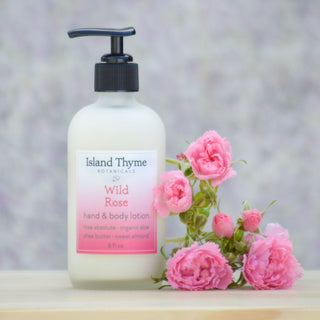 A bottle of Wild Rose Lotion rests beside pink roses on a light wooden surface, infusing the air with a natural rose scent and set against a gently blurred background.