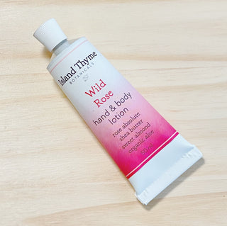A tube of Wild Rose Lotion, infused with rose absolute, shea butter, sweet almond, and organic aloe for a natural rose scent, sits on a light wood surface.