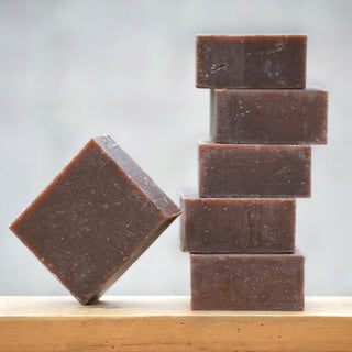 Six Cinnamon Clove Soap bars, rectangular and brown, are arranged on a wooden surface—five stacked vertically and one leaning diagonally. Infused with cinnamon bark essential oils, they exude a warm spicy scent. The background is plain and blurred.