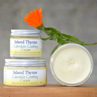 Two jars of Calendula Comfrey Cream, an all-natural moisturizer for dry skin, are stacked with one open to show the creamy contents. An orange calendula flower rests on top against a neutral background.