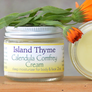 A 2 oz glass jar of Calendula Comfrey Cream with a silver lid rests on wood beside an orange calendula flower. This all-natural deep dry skin cream is for body and face.