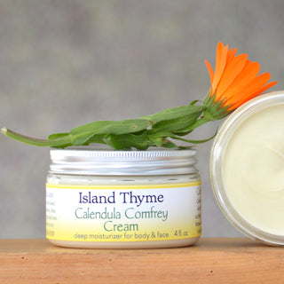 A 4 fl oz jar of Calendula Comfrey Cream with its lid off reveals the rich cream inside, topped with a calendula flower. This all-natural moisturizer is ideal for body and face, perfect as a dry skin cream.