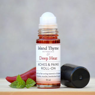 A Deep Heat Liquid Roll-On stands upright with its cap off, accompanied by a red chili pepper and fresh mint leaves on a light wooden surface.