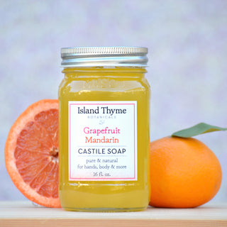 A mason jar labeled Grapefruit Mandarin Castile Soap sits between a halved grapefruit and a whole mandarin orange with a leaf, set against a soft pastel background.
