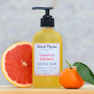 A bottle of Grapefruit Mandarin Castile Soap, crafted with essential oils, is placed between half a grapefruit and a whole mandarin with a leaf, set against a softly blurred background.