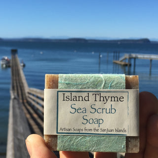 A hand holds a bar of Sea Scrub Soap with a dock and calm blue water in the background under a clear sky. The label reads Artisan Soaps from the San Juan Islands.