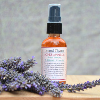A bottle of Aches & Pains Oil with Arnica Montana, St. John’s Wort, and Lavender sits on a wooden surface, surrounded by fresh lavender sprigs. The pump-top bottle features a clear label.