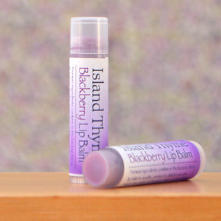 Two tubes of Blackberry Lip Balm with purple labels—one standing upright and one lying down—rest on a wooden surface, set against a softly blurred background that highlights their moisturizing blackberry formula.