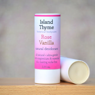 A tube of Rose Vanilla Deodorant stands upright, while another lies on its side. The pink and white label highlights its biodegradable packaging and lists rose absolute as a key ingredient.