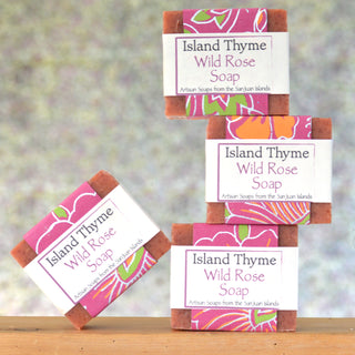 Four bars of Wild Rose Soap, enriched with rosehips, are stacked on a wooden surface. Their colorful wrappers read: “Artisan Soaps from the San Juan Islands” against a blurred pastel background.
