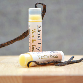Two tubes of Vanilla Lip Balm, crafted with natural, gluten-free ingredients, are shown on a wooden surface with vanilla bean pods, set against a neutral gray background.