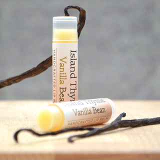 Two tubes of Vanilla Lip Balm made with natural ingredients are displayed on a wooden surface beside vanilla bean pods. One stands upright and the other lies in front, highlighting this gluten-free, handcrafted favorite.