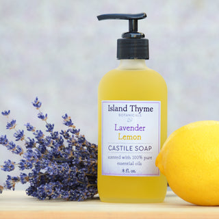 A bottle of Lavender Lemon Castile Soap is placed between dried lavender and a fresh lemon on a light surface with a soft, blurred background.