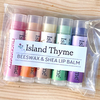 A clear bag holds the Lip Balm Classic Set—six colorful Island Thyme Lip Balm tubes, each with a distinct flavor label—arranged on a light wooden surface.