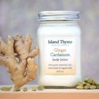 A jar of Ginger Cardamom Lotion rests on a surface with fresh ginger root and cardamom pods. Its label features essential oils, shea butter, and organic aloe—ideal for nourishing dry skin.