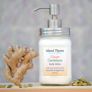 A glass pump jar of Ginger Cardamom Lotion rests on a wooden surface beside fresh ginger root and cardamom pods, with a softly blurred background—the perfect treat for dry skin.