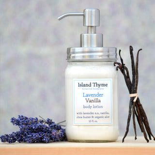 A bottle of Lavender Vanilla Lotion, featuring shea butter and a pump dispenser, sits beside lavender flowers and vanilla beans against a softly blurred backdrop.