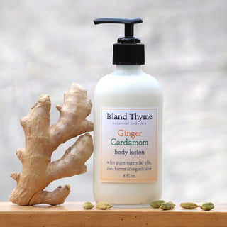 A bottle of Ginger Cardamom Lotion for dry skin sits beside fresh ginger root and green cardamom pods on a wooden surface. This nourishing blend uses essential oils to help leave skin smooth and hydrated.