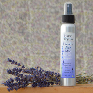 A silver spray bottle labeled Lavender Aloe Mist 4oz, made with aloe vera and lavender essential oil, sits next to dried lavender on a wooden surface against a blurred, textured background.