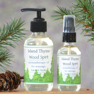 Two bottles of Wood Spirit Aromatherapy Massage Oil, one pump (4 oz) and one spray (2 oz), rest beside pine branches and a pine cone. Labels show use as bath, body massage oil, and natural moisturizer.