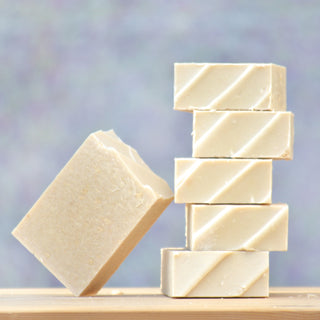 Six Naked Goat Soap bars for sensitive skin are arranged with five stacked vertically and one leaning diagonally, set against a soft, pastel-toned background.