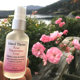 A hand holds a bottle of Rose Rejuvenation Toner near blooming pink roses, with a wooden bench, lake, and forested hills in the background—capturing a serene moment of balancing skin pH surrounded by nature’s beauty.