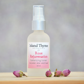 A frosted glass bottle of Rose Rejuvenation Toner with a spray cap, made from natural ingredients to balance skin pH, is surrounded by dried rosebuds on a wooden surface against a softly blurred background.