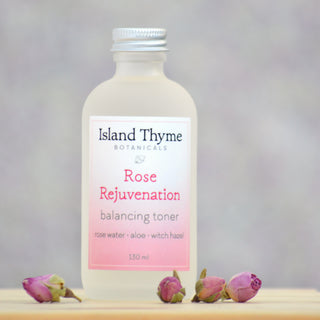 A 130 ml frosted glass bottle of Rose Rejuvenation Toner stands on a light surface amid dried rosebuds. Made with natural ingredients, it helps balance skin pH for a refreshed, radiant complexion.