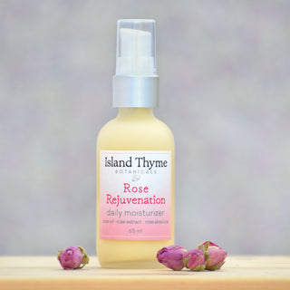 A bottle of Rose Rejuvenation Daily Moisturizer rests on a light surface beside three dried rosebuds, with a softly blurred pastel background highlighting its elegance.