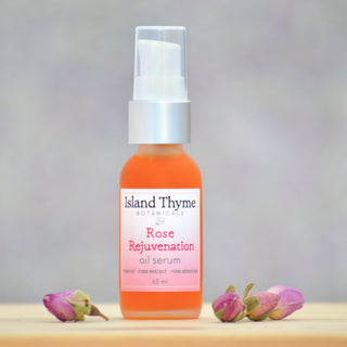 A bottle of Rose Rejuvenation Facial Oil Serum sits on a light surface between two dried rosebuds, its antioxidant-rich formula full of essential fatty acids. The softly blurred background gives a gentle, serene feel.