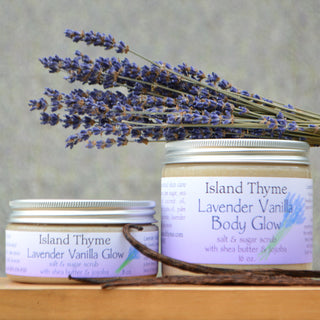 Two jars of Lavender Vanilla Glow Salt & Sugar Scrub exfoliate and moisturize, displayed with a bundle of dried lavender on a wooden surface against a neutral background.