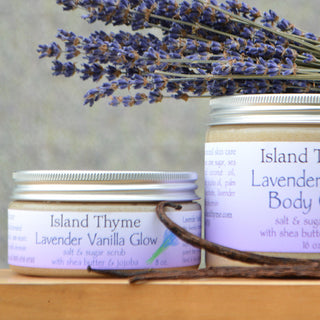 A jar and tin of Island Thymes Lavender Vanilla Glow Salt & Sugar Scrub sit in front, with dried lavender in the background. Labels highlight its gentle exfoliation and moisturizing benefits for radiant, silky-soft skin.
