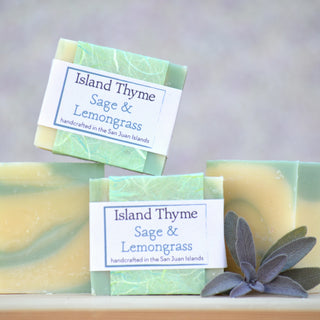 Three bars of Sage & Lemongrass Soap made with pure essential oils are displayed on a light surface—two stacked, one upright—with a sage leaf beside them and a softly blurred background.