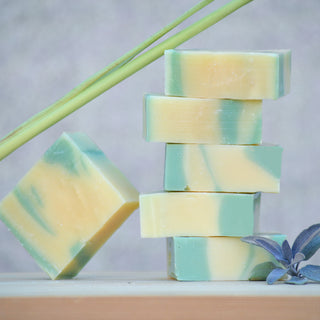 Six Sage & Lemongrass Soap bars, marbled yellow and green, are stacked on wood with one bar leaning. Infused with lemongrass essential oil, the display includes a lemongrass stalk and sage sprig against a soft gray background.