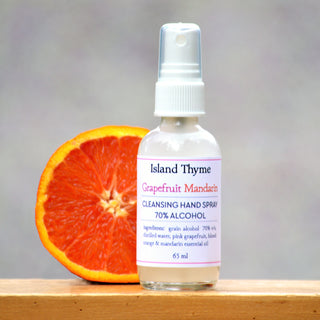 A bottle of Grapefruit Mandarin 70% Alcohol Hand Cleansing Spray sits beside a sliced grapefruit on a wooden surface, its label showing 70% alcohol and essential oils, all set against a neutral background.