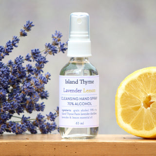 A bottle of Lavender Lemon 70% Alcohol Hand Cleansing Spray rests on a wooden surface, next to a sprig of lavender and a sliced lemon.