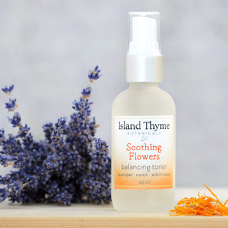 A bottle of Soothing Flowers Balancing Toner, formulated with lavender, neroli, and witch hazel to rejuvenate skin and support natural pH, is displayed on wood beside purple lavender and orange flower petals.