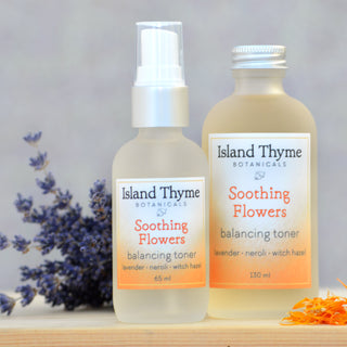 Two bottles of Soothing Flowers Balancing Toner are displayed on a light surface—one with a spray top, one with a screw cap. Lavender flowers are arranged nearby for decoration.
