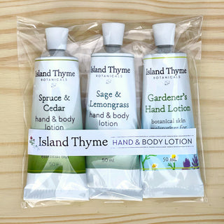 Three tubes of Island Thyme’s Forest & Garden Travel Lotion Trio—Spruce & Cedar, Sage & Lemongrass, and Gardener’s Hand Lotion—stand side by side on a light wood background, packaged together with a clear biodegradable band.