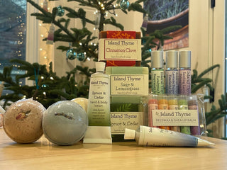 A variety of Island Thyme bath and body products, including soaps, lotions, lip balms, and bath bombs, are displayed on a wooden table in front of a decorated Christmas tree.