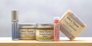 Five Island Thyme products lined up on a wooden surface: a roll-on stick, two jars, a lip balm, and a bar of Orcas Lavender handcrafted soap against a light background.
